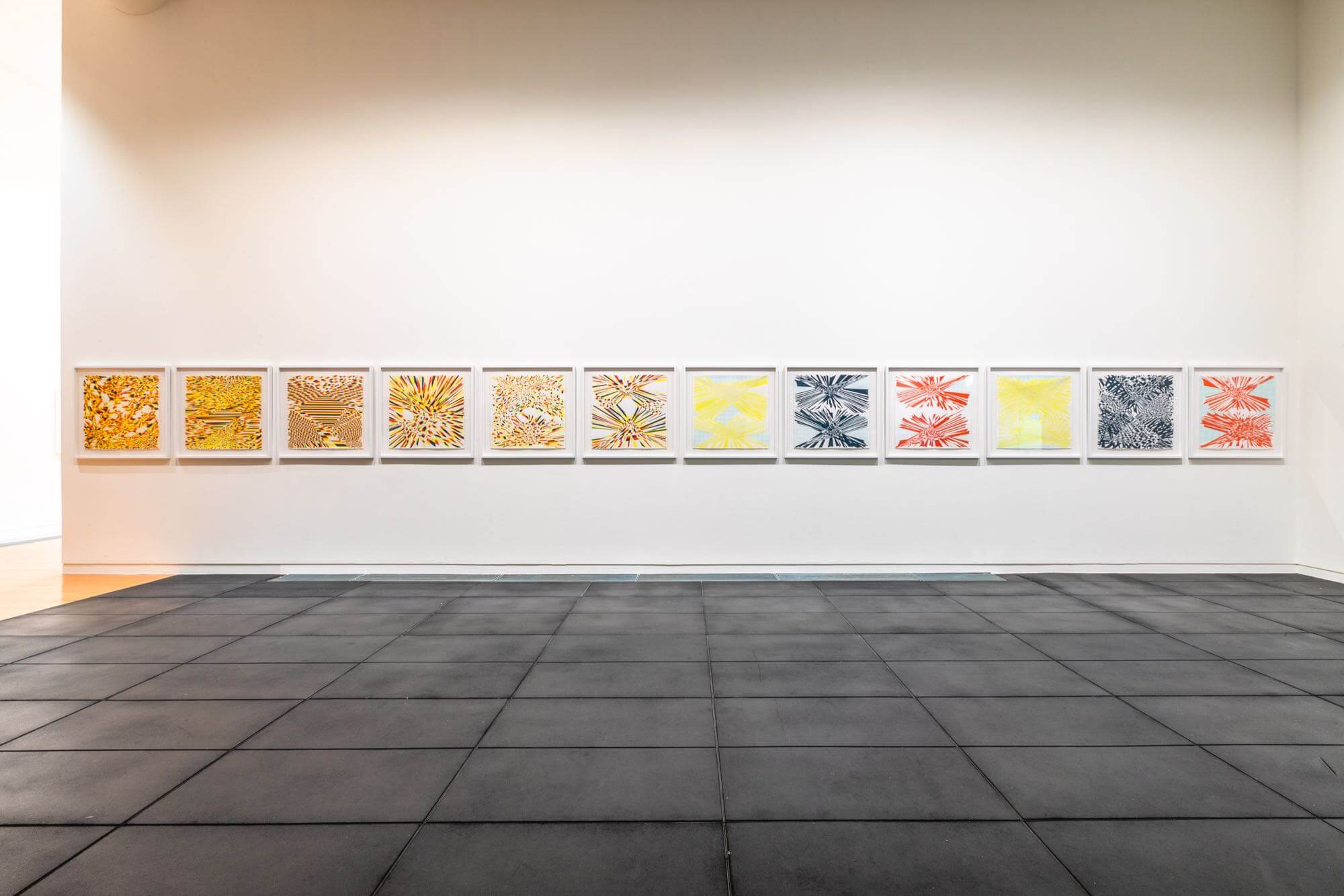 Installation view of twelve watercolour works in a long line. The receding floor is black square tiled rubber, the wall white.